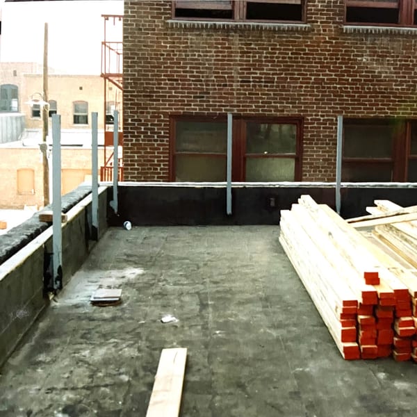 Rooftop under construction with stacks of lumber, metal posts, and a brick building in the background.